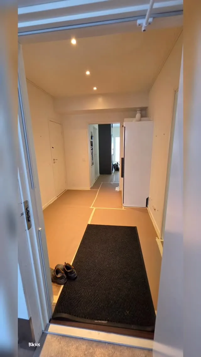 Hall i lägenhet med golv täckt av skyddspapp, tejpade skarvar, dörrmatta, vita väggar och spotlights i taket