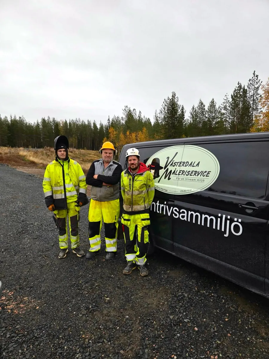 Tre arbetare i varselkläder står vid en svart skåpbil med Västerdala Måleriservice logga på en grusplan vid skogen